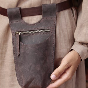 May include: A brown leather pouch with a zipper closure and a small pocket on the front. The pouch is attached to a brown leather belt.