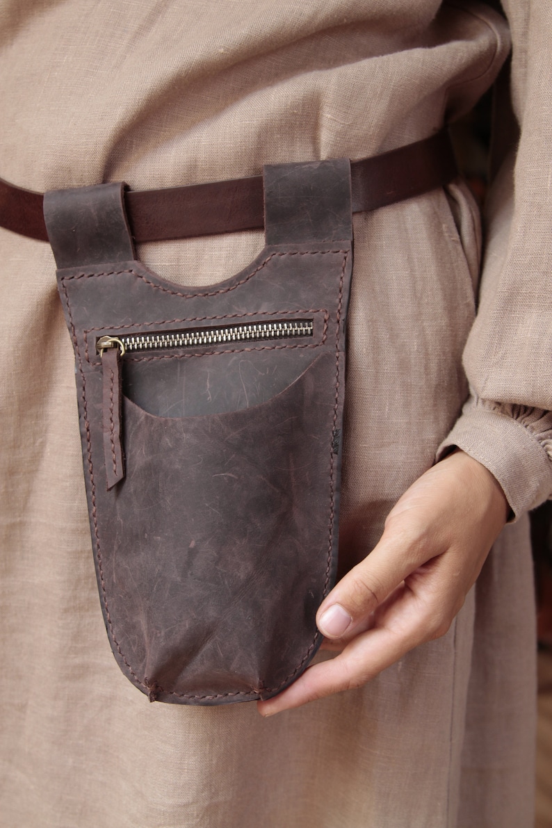 May include: A brown leather pouch with a zipper closure and a small pocket on the front. The pouch is attached to a brown leather belt.