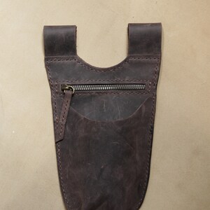 May include: A brown leather pouch with a zippered pocket and a flap closure. The pouch is designed to be worn on a belt or strap.