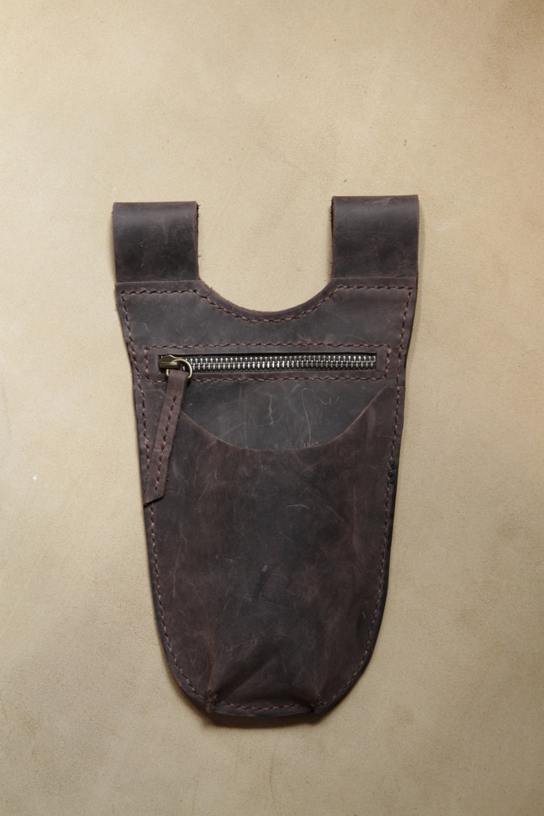 May include: A brown leather pouch with a zippered pocket and a flap closure. The pouch is designed to be worn on a belt or strap.