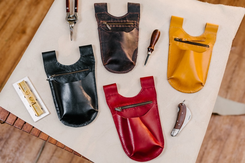 May include: Four leather pouches with zippered pockets and a single open pocket. The pouches are in brown, black, yellow, and red. The pouches are on a white surface with a wooden surface in the background.