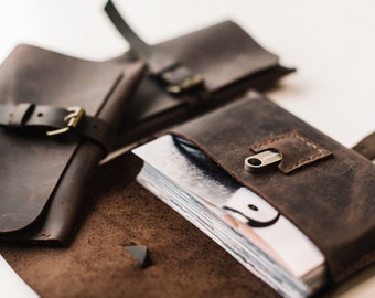 May include: A brown leather journal with a strap and buckle closure. The journal is open to reveal a stack of white pages. A small silver USB drive is tucked into a pocket on the cover.