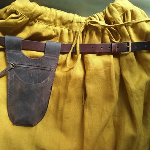 May include: A brown leather pouch with a zipper and a pocket hangs from a brown leather belt with a gold buckle. The belt is worn over a yellow linen skirt.