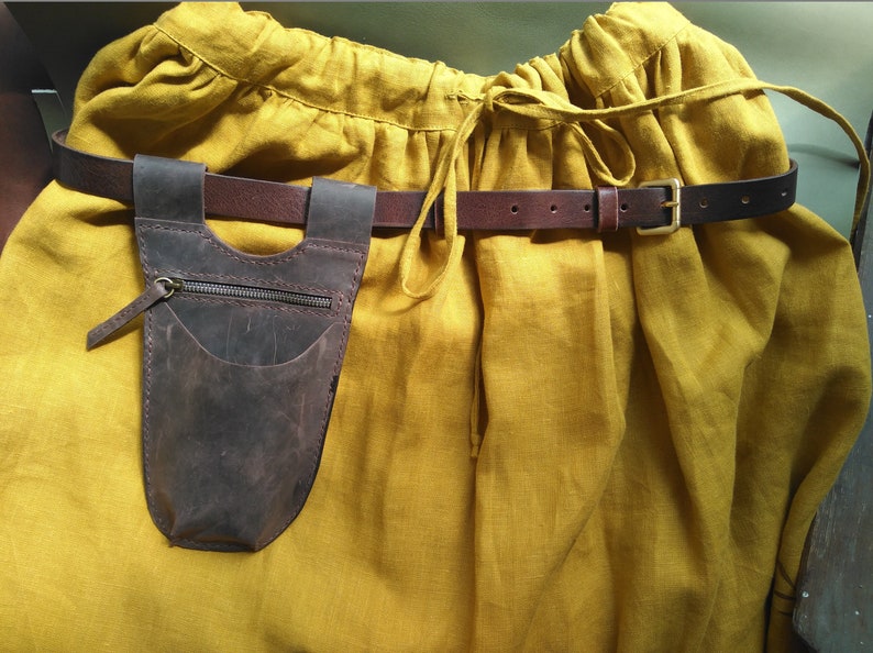 May include: A brown leather pouch with a zipper and a pocket hangs from a brown leather belt with a gold buckle. The belt is worn over a yellow linen skirt.
