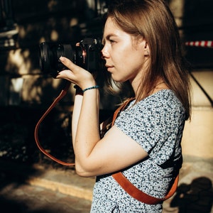 May include: A woman in a blue floral dress with a brown camera strap is holding a black camera with a lens and taking a photo.
