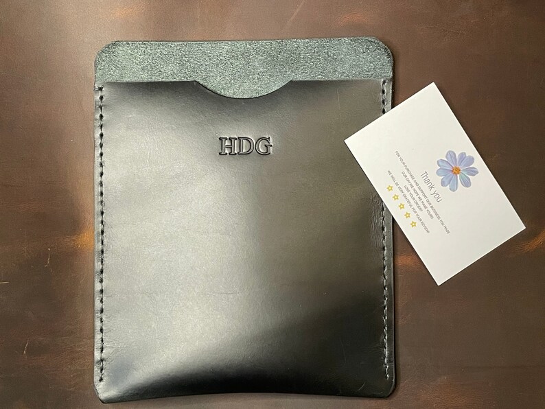 May include: Black leather wallet with a stitched edge and the initials "HDG" embossed on the front. A small white card with a blue flower and the text "Thank You" is next to the wallet.