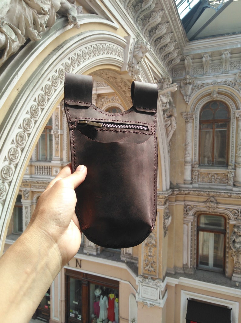 May include: A brown leather pouch with a zippered pocket and a flap closure. The pouch is held in a hand against a background of an ornate building.