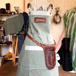 Puede incluir: Un delantal verde salvia con un panel superior gris claro y correas de cuero marr&oacute;n. Un parche rectangular de cuero marr&oacute;n con el texto MON BUREAU est&aacute; centrado en el pecho. Una funda de cuero marr&oacute;n con bolsillo con cremallera est&aacute; en la cintura.