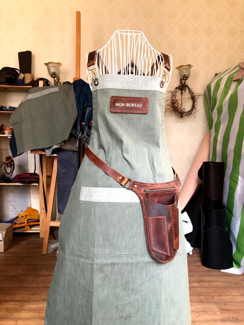Puede incluir: Un delantal verde salvia con un panel superior gris claro y correas de cuero marr&oacute;n. Un parche rectangular de cuero marr&oacute;n con el texto MON BUREAU est&aacute; centrado en el pecho. Una funda de cuero marr&oacute;n con bolsillo con cremallera est&aacute; en la cintura.