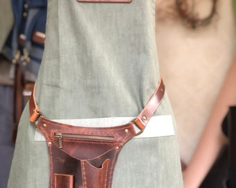 May include: A sage green canvas apron featuring a brown leather utility belt attachment. The apron has a rectangular leather patch with the text MON BUREAU embossed in white. The leather belt includes a zippered pocket and two open tool slots.