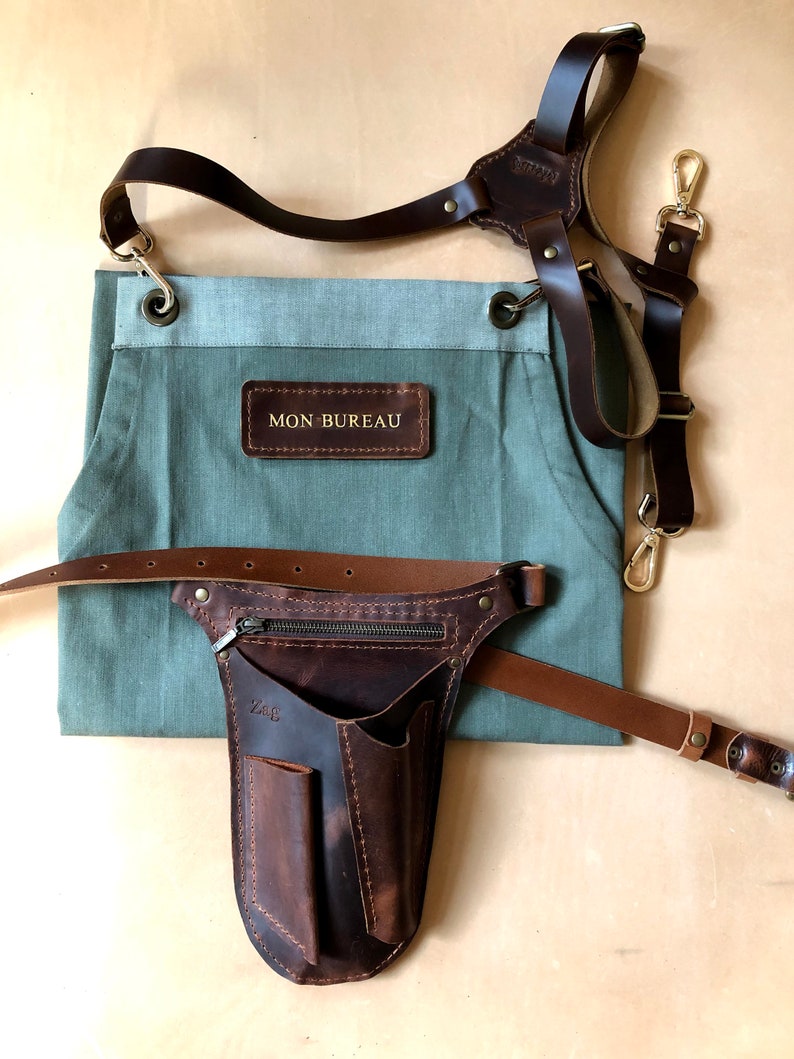 Puede incluir: Un delantal verde salvia con un parche de cuero marr&oacute;n con el texto MON BUREAU, combinado con correas de hombro de cuero marr&oacute;n oscuro y una funda de herramientas de cuero marr&oacute;n con bolsillo con cremallera y el texto ZAG.