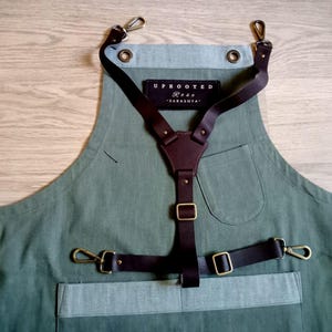 Puede incluir: Un delantal verde oliva con un sistema de arn&eacute;s de cuero marr&oacute;n oscuro y herrajes de tono lat&oacute;n. Un parche rectangular negro muestra el texto blanco UPROOTED Rose SARASOTA. El delantal incluye un bolsillo en el pecho, uno en la cintura y ojales.