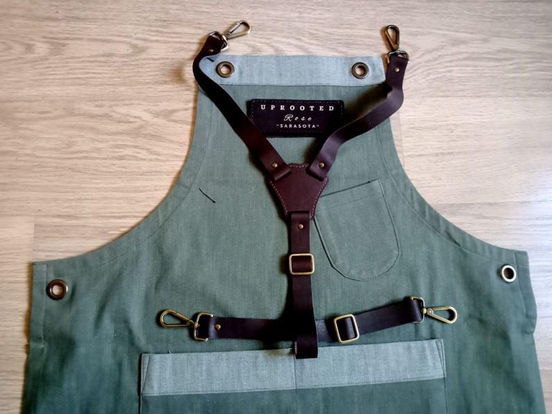 Puede incluir: Un delantal verde oliva con un sistema de arn&eacute;s de cuero marr&oacute;n oscuro y herrajes de tono lat&oacute;n. Un parche rectangular negro muestra el texto blanco UPROOTED Rose SARASOTA. El delantal incluye un bolsillo en el pecho, uno en la cintura y ojales.