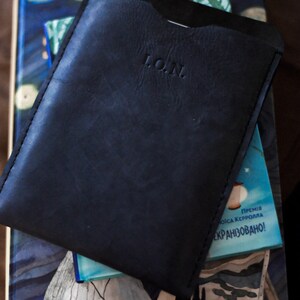 May include: A black leather phone case with the word "L.O.N." embossed on the front. The case is sitting on top of a book with a blue and white cover.