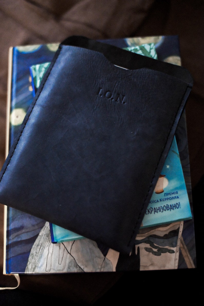 May include: A black leather phone case with the word "L.O.N." embossed on the front. The case is sitting on top of a book with a blue and white cover.