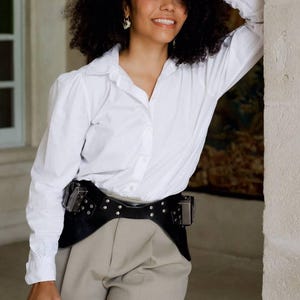 Puede incluir: Una persona con una camisa blanca abotonada, pantalones color beige y un cinturón de cuero negro con tachuelas plateadas y dos bolsillos. La persona sostiene una cámara negra con una lente.