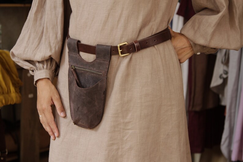 May include: A brown leather belt with a brown leather pouch attached. The pouch has a zipper and a small pocket. The belt is worn over a light brown linen dress.
