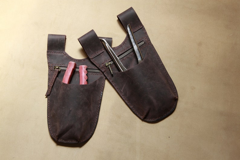 May include: Two brown leather tool pouches with zippered pockets. One pouch has a pair of pink pliers inside, and the other has a pair of metal tweezers.