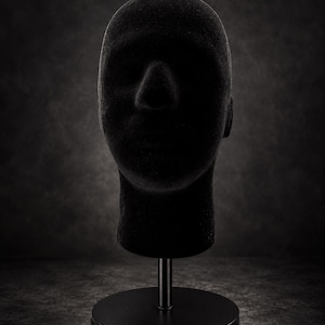 Puede incluir: Un maniquí de cabeza negro mate con una superficie texturizada similar al terciopelo. La cabeza está montada sobre una varilla vertical simple y delgada unida a una base de metal negro circular y plana para exhibir accesorios.
