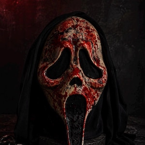 Könnte beinhalten: Eine weiße geisterhafte Maske mit einer abgenutzten, rissigen Textur und intensiven roten Blutspritzern. Die Maske hat große, hohle schwarze Augenhöhlen und einen länglichen, offenen schwarzen Mund, befestigt an einer schwarzen Stoffkapuze.