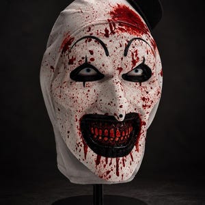 Puede incluir: Una máscara de payaso blanca con un pequeño sombrero de copa negro, maquillaje de ojos negro y una amplia sonrisa con dientes. La máscara está cubierta de salpicaduras rojas similares a sangre por toda la cara y la frente.