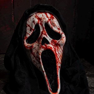 Op de afbeelding: Een wit spookachtig masker met een lang, vervormd gezicht, grote zwarte oogkassen en een langwerpige open mond. Het masker is bedekt met bloedrode spetters en is bevestigd aan een zwarte stoffen kap die rond de zijkanten en bovenkant valt.