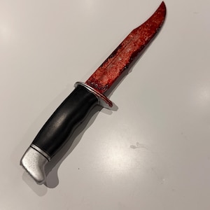 Può includere: Un coltello a lama fissa con manico nero, pomolo e guardia argentati. La lama è ricoperta da una finitura rossa brillante e testurizzata che imita l'aspetto di schizzi di sangue, estendendosi dalla base fino alla punta affilata.