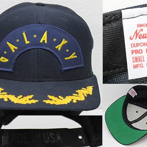 May include: A navy blue baseball cap with a yellow embroidered design of a star and the word "GALAXY" on the front. The cap has a green underside and a black label on the back that reads "Since 1920 New Era DUPONT VISOR PRO MODEL SMALL - MEDIUM MFG. IN U.S.A."