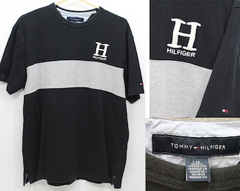 May include: Black and grey striped t-shirt with a white Tommy Hilfiger logo on the front. The logo features the letter "H" and the text "Hilfiger".