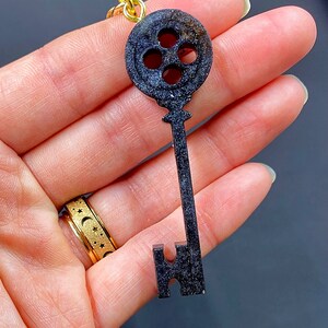 Puede incluir: Encanto de llave negro en forma de llave con una cabeza circular que presenta cuatro agujeros. El encanto tiene un acabado brillante y brillante.