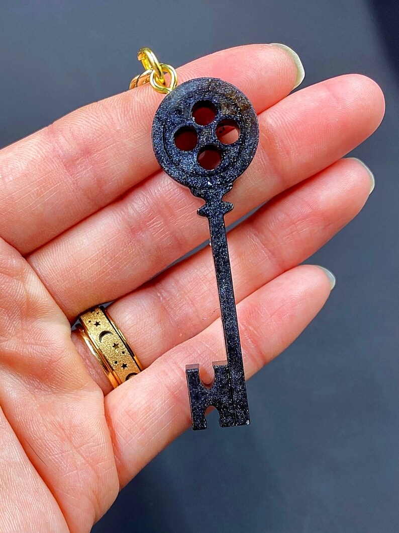 Puede incluir: Encanto de llave negro en forma de llave con una cabeza circular que presenta cuatro agujeros. El encanto tiene un acabado brillante y brillante.