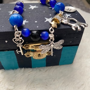 May include: A blue and silver charm bracelet with a dragonfly, key, and star charms. The bracelet is on a black and teal striped box with white stars.