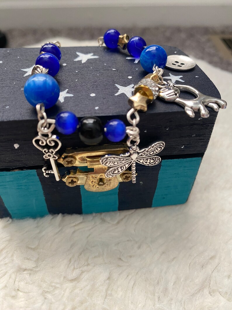 May include: A blue and silver charm bracelet with a dragonfly, key, and star charms. The bracelet is on a black and teal striped box with white stars.