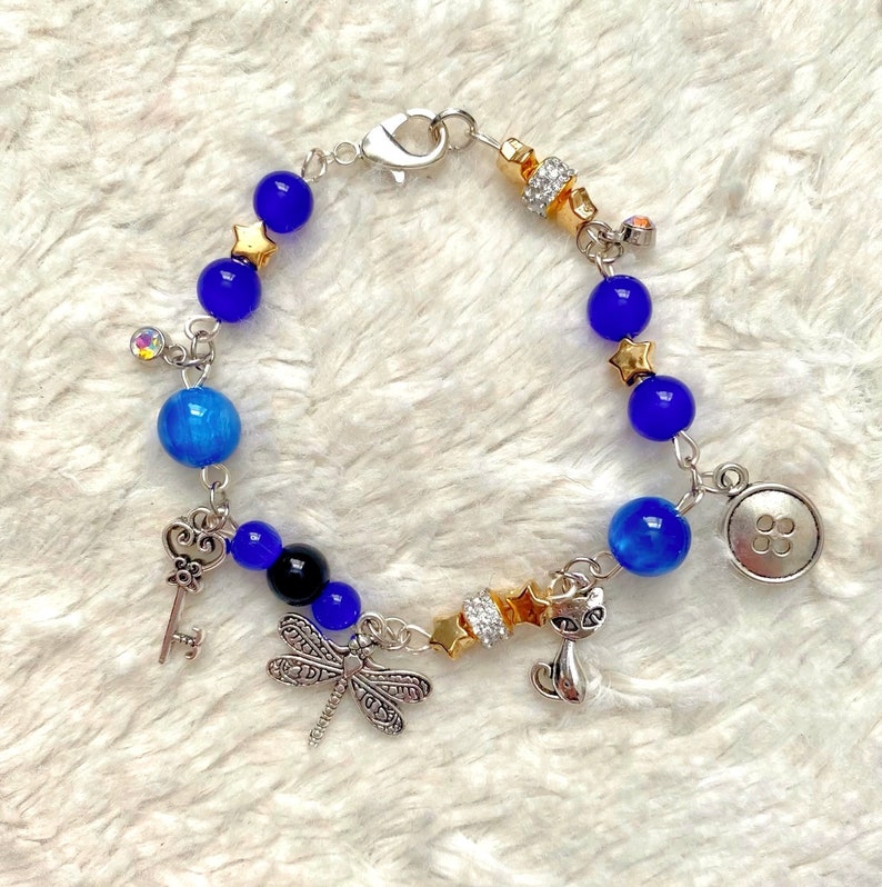 May include: A blue and silver charm bracelet with a variety of charms, including a key, a dragonfly, a cat, a button, and stars.