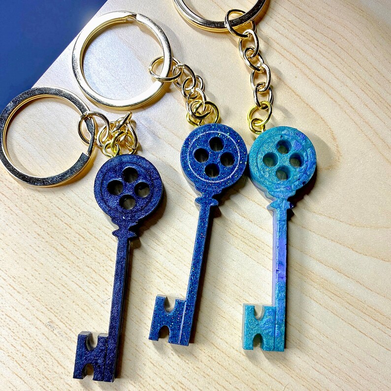 Puede incluir: Tres llaveros con dijes de llave de resina azul y verde en forma de llaves con un dise&ntilde;o de bot&oacute;n. Los dijes est&aacute;n unidos a llaveros de metal dorado.