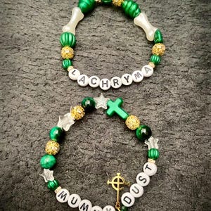 May include: Two beaded bracelets with white letter beads spelling "LACHRYMA" and "MUMMY DUST". The bracelets feature green, gold, and white beads, with cross and star charms. One bracelet has bone-shaped beads.