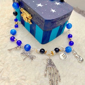May include: A silver chain necklace with blue and black beads, a silver skeleton hand charm, a dragonfly charm, a key charm, a button charm, and a silver teardrop charm. The necklace is displayed on a blue and white wooden box with stars.