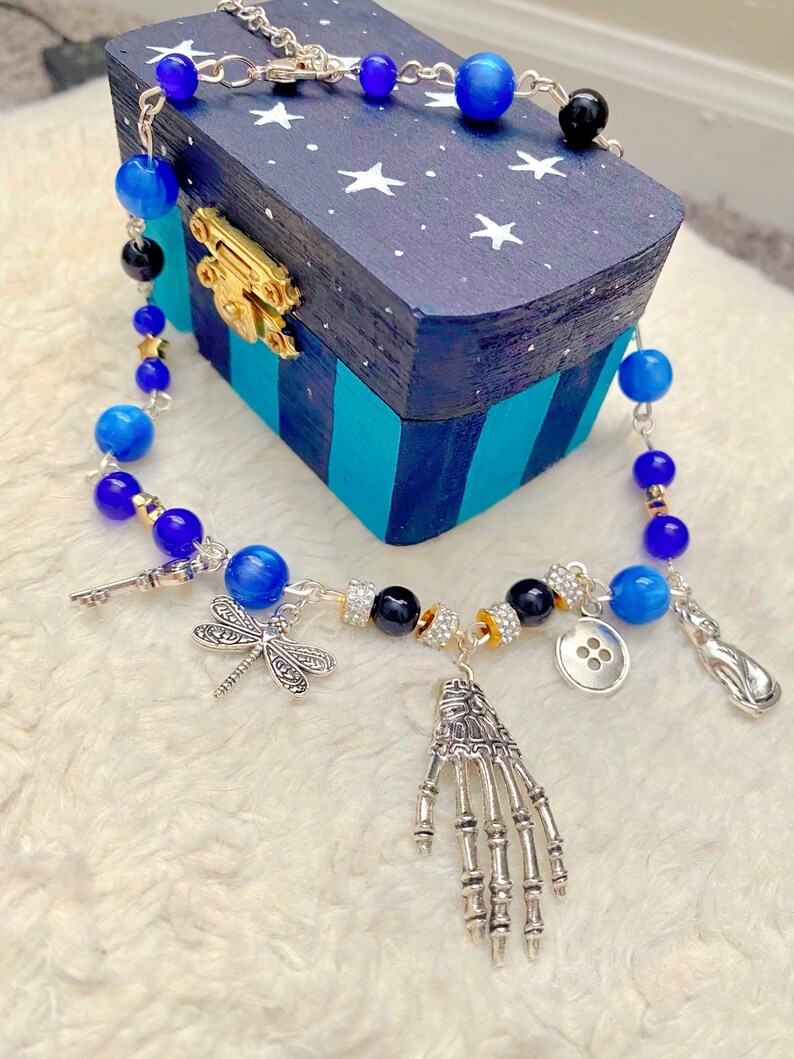 May include: A silver chain necklace with blue and black beads, a silver skeleton hand charm, a dragonfly charm, a key charm, a button charm, and a silver teardrop charm. The necklace is displayed on a blue and white wooden box with stars.
