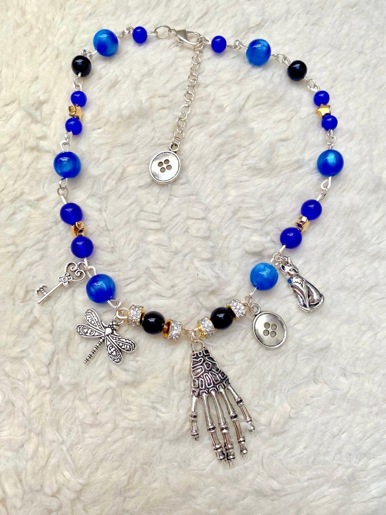 May include: A blue and silver necklace with a skeleton hand charm, dragonfly charm, key charm, and two buttons. The necklace also features blue and black beads, gold stars, and clear rhinestones.
