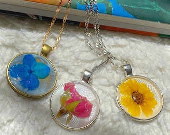 May include: Three necklaces with different flower pendants. The pendants are round and have a clear resin covering. The flowers are a blue hydrangea, a pink rose, and a yellow sunflower.