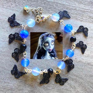 Puede incluir: Una pulsera plateada con cuentas azules y negras, y dijes de mariposa negros. La pulsera tiene una pequeña imagen de un personaje de dibujos animados en el centro. La pulsera está diseñada con una cadena delicada.