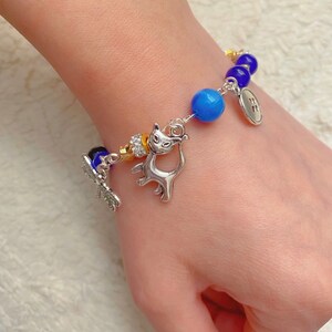 May include: A silver charm bracelet with blue and purple glass beads, a silver cat charm, a silver dragonfly charm, and a silver button charm.