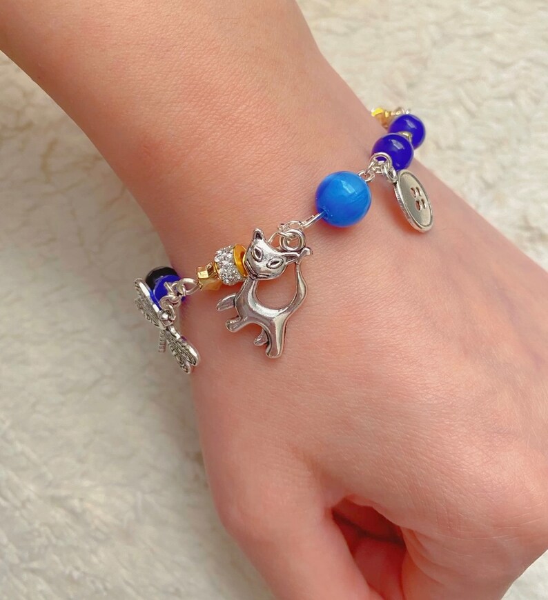 May include: A silver charm bracelet with blue and purple glass beads, a silver cat charm, a silver dragonfly charm, and a silver button charm.