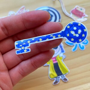 Puede incluir: Una pegatina en forma de llave azul y amarilla con estrellas y un extremo en forma de globo. La pegatina est&aacute; sobre una superficie de madera.