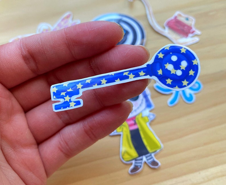Puede incluir: Una pegatina en forma de llave azul y amarilla con estrellas y un extremo en forma de globo. La pegatina est&aacute; sobre una superficie de madera.