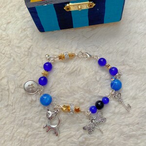 May include: A silver charm bracelet with blue and black beads, a silver cat charm, a dragonfly charm, a key charm, and a button charm. The bracelet is on a white fuzzy surface.