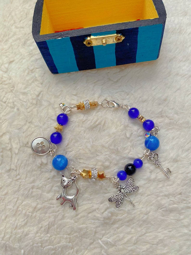 May include: A silver charm bracelet with blue and black beads, a silver cat charm, a dragonfly charm, a key charm, and a button charm. The bracelet is on a white fuzzy surface.