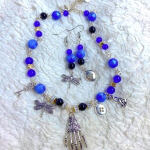 May include: A blue and silver necklace with a skeleton hand charm, dragonfly charms, and buttons. Matching blue and silver earrings with dragonfly charms.