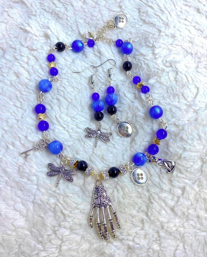 May include: A blue and silver necklace with a skeleton hand charm, dragonfly charms, and buttons. Matching blue and silver earrings with dragonfly charms.