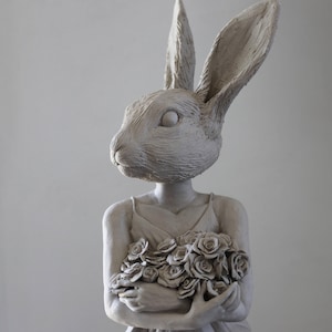 Könnte beinhalten: Eine hellgraue Skulptur einer Figur mit einem Kaninchenkopf, die ein Kleid trägt und einen Rosenstrauß hält. Die Skulptur hat eine strukturierte Oberfläche und steht vor einem schlichten Hintergrund.
